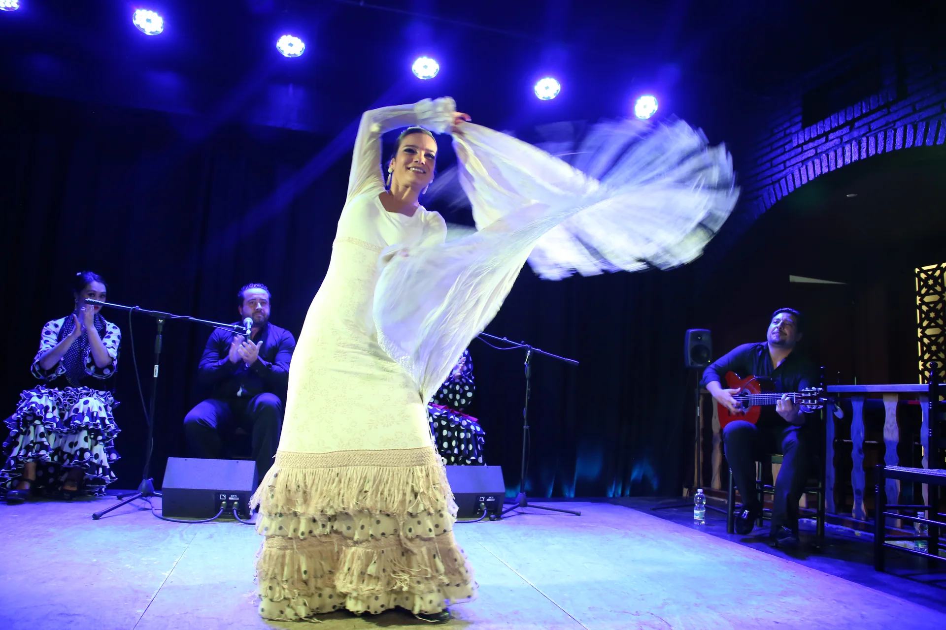 A flamenco dancer mid-turn on a tablao stage, cuadro of singer and guitarist either side
